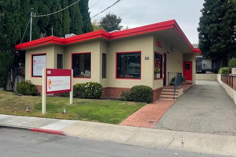 The California Hearing Center office at 88 N. San Mateo Drive seen from across the street: a long single-storey building with a bright red roofline, a lawn, a brick path to the front steps, and the driveway running down its right-hand side.